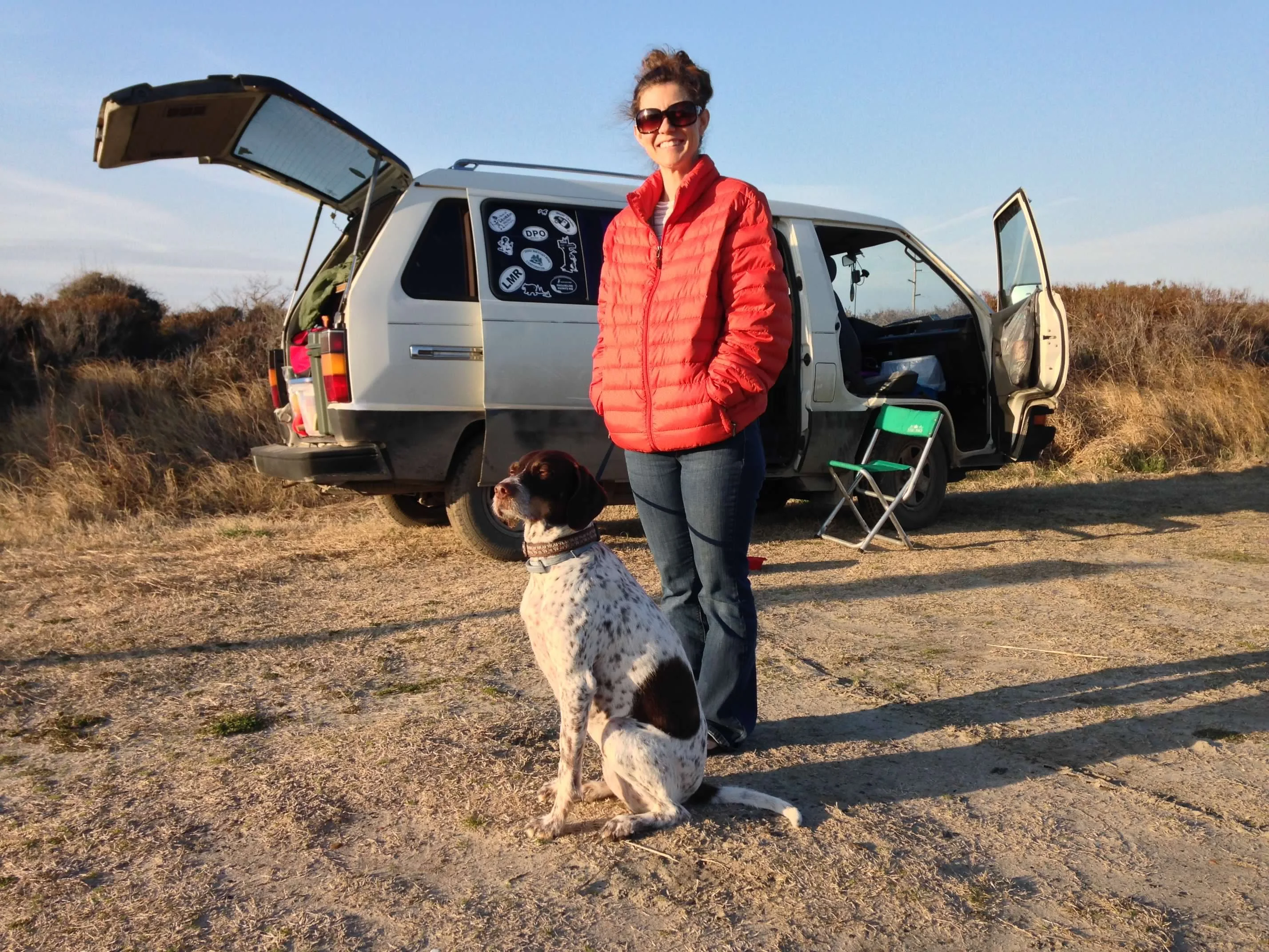 Polly and the dog standing next to the van parked among sand dunes and beach grass